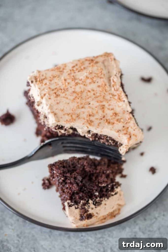 Homemade chocolate cake is soaked with Irish cream and topped with whipped cream frosting and a sprinkle of chocolate shavings for a decadent and creamy Irish Cream Poke Cake. A slice of Irish Cream Poke Cake with a bite taken out, highlighting the layers of cake, Irish cream, and whipped cream