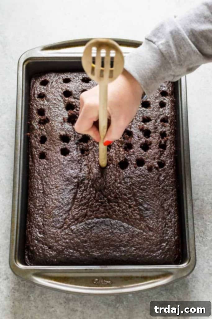 Homemade chocolate cake is soaked with Irish cream and topped with whipped cream frosting and a sprinkle of chocolate shavings for a decadent and creamy Irish Cream Poke Cake. Using the end of a wooden spoon to poke uniform holes into a freshly baked chocolate cake, ready for liquid infusion