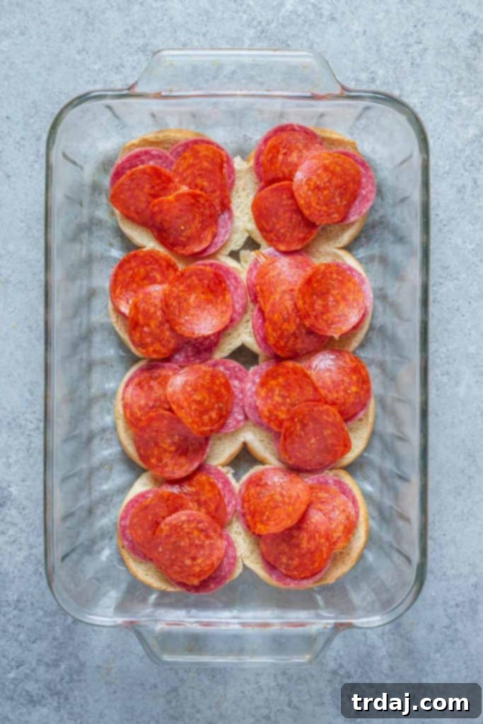Salami slices layered on the bottom half of slider rolls, forming the first layer of the sandwich.