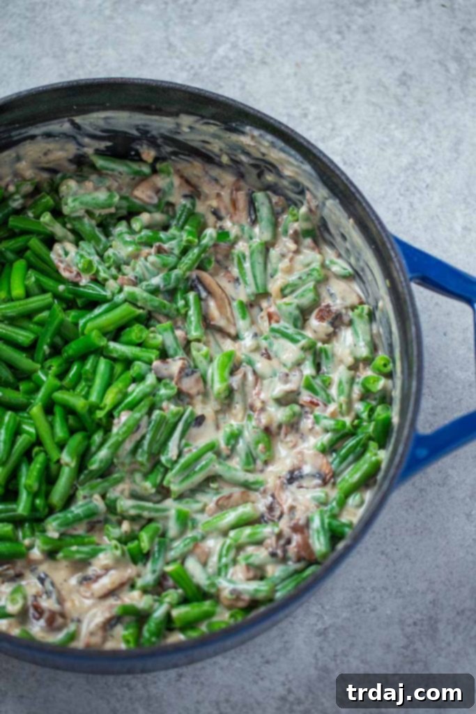 Cooked green beans being added and folded into the homemade cream of mushroom soup in a skillet, preparing for the casserole.
