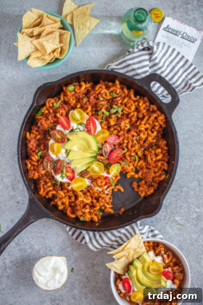 Close-up of Healthier One Pan Taco Pasta in a rustic cast iron pan, garnished with sour cream, diced tomatoes, and avocado, with a single serving bowl visible in the foreground