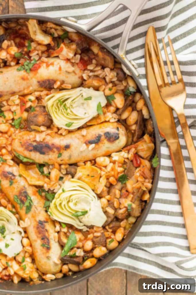 Close-up of golden brown Italian sausages nestled in a rich tomato sauce with farro, cannellini beans, and artichokes in a skillet.