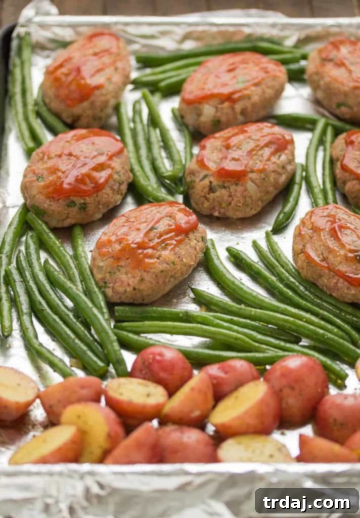 Sheet Pan Mini Meatloaves with Potatoes and Green Beans: Prep your effortless meal on one pan before baking to perfection. An unbaked sheet pan dinner featuring mini meatloaves topped with ketchup, surrounded by seasoned potatoes and fresh green beans, ready for the oven.