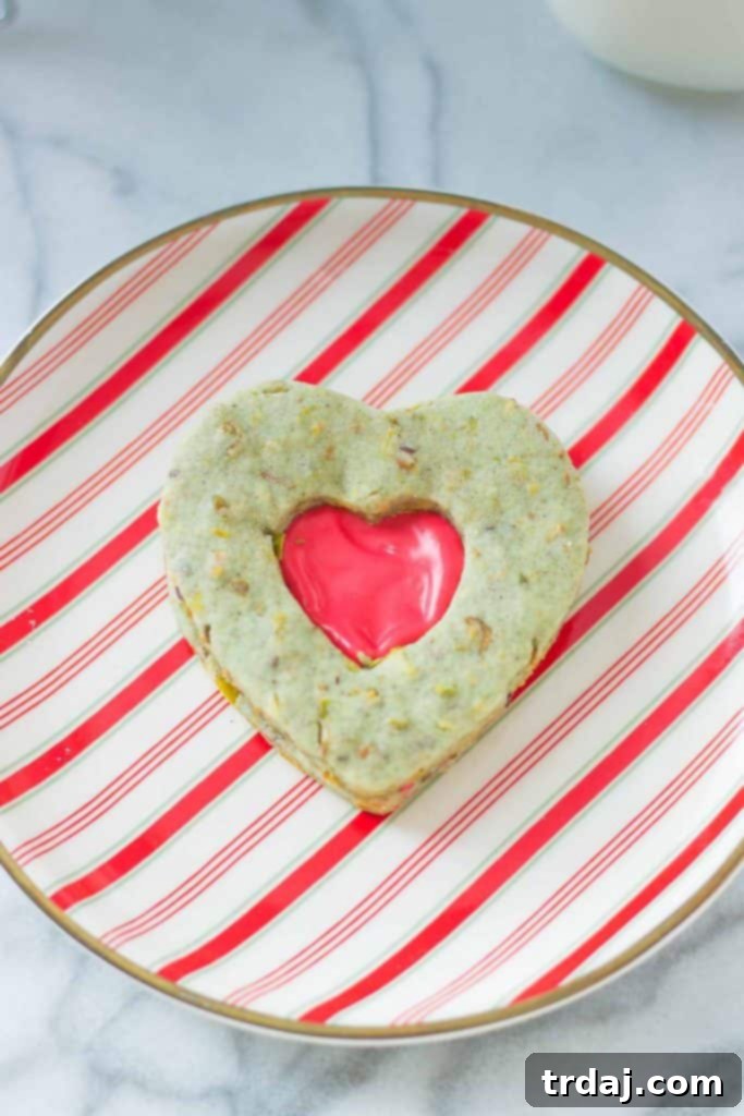 Close-up view of a Grinch Sandwich Cookie, highlighting the rich green pistachio shortbread and the vibrant red marshmallow filling visible through the heart-shaped cutout. A delightful Christmas cookie recipe.
